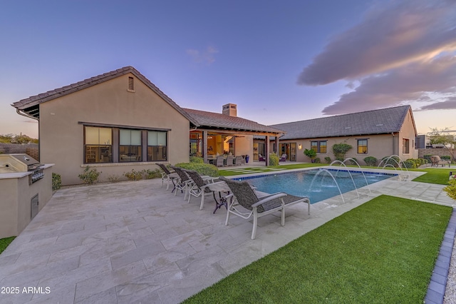 pool at dusk featuring a lawn, an outdoor kitchen, a grill, pool water feature, and a patio