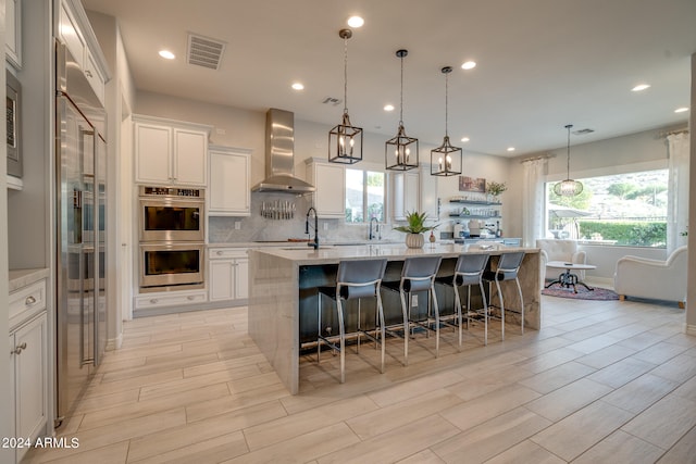 kitchen with wall chimney range hood, white cabinets, an island with sink, and light hardwood / wood-style flooring