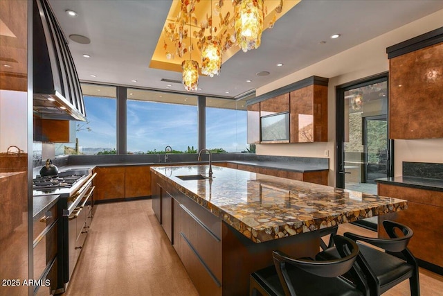 kitchen featuring a kitchen island with sink, wall chimney exhaust hood, brown cabinetry, gas stovetop, and a sink