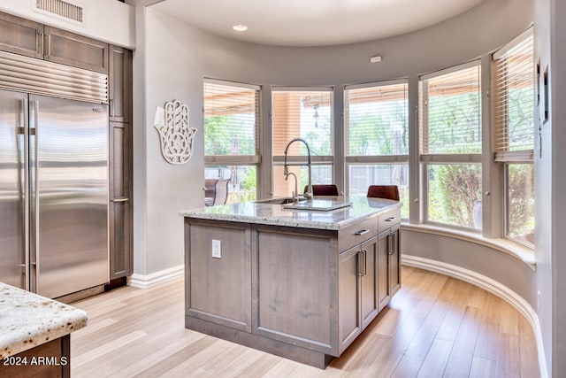 kitchen featuring light wood-type flooring, built in fridge, a kitchen island with sink, and light stone countertops