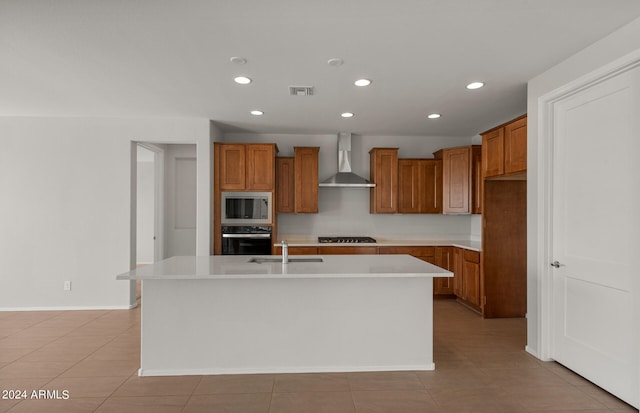 kitchen with wall chimney range hood, light tile patterned floors, appliances with stainless steel finishes, an island with sink, and sink