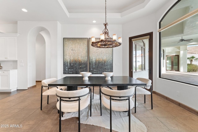 tiled dining room with ceiling fan with notable chandelier and a tray ceiling