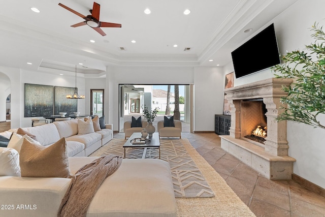 living room featuring a raised ceiling, ceiling fan, a fireplace, and crown molding