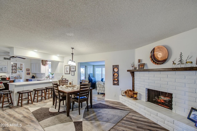 dining space featuring a brick fireplace, a textured ceiling, light wood-type flooring, and ceiling fan