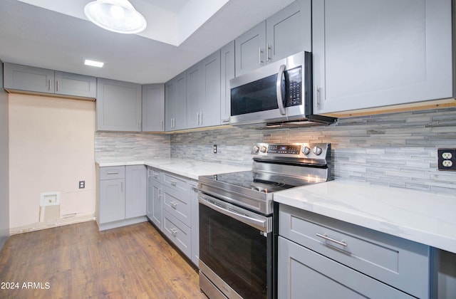 kitchen featuring appliances with stainless steel finishes, wood-type flooring, gray cabinetry, decorative backsplash, and light stone counters