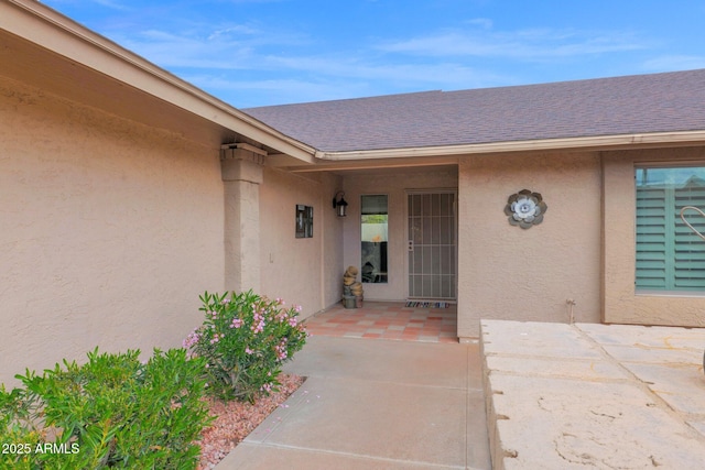 view of exterior entry featuring stucco siding, roof with shingles, and a patio area