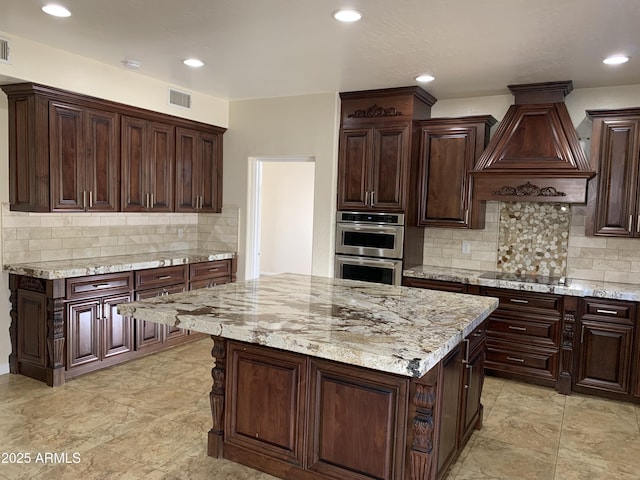 kitchen featuring double oven, light stone counters, a kitchen island, visible vents, and custom range hood