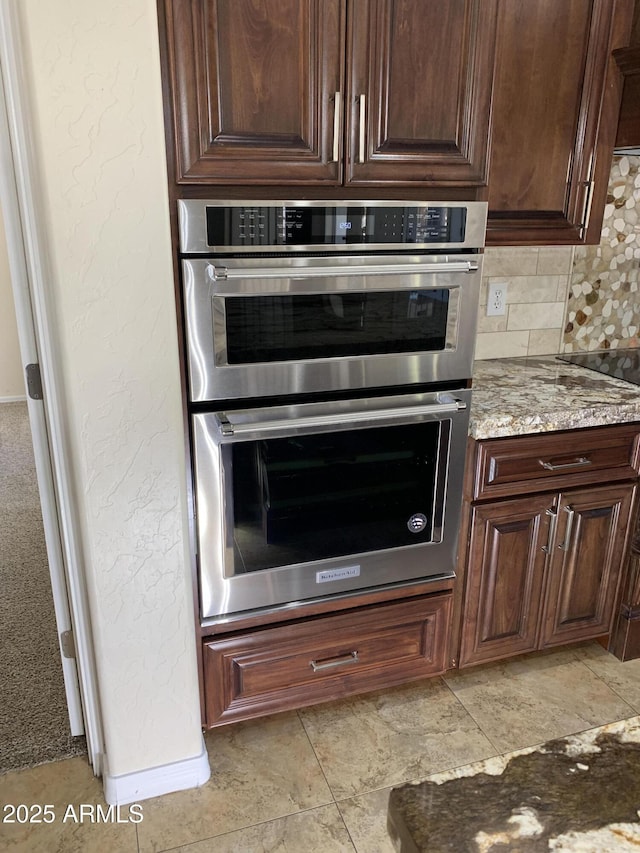 kitchen with a textured wall, double oven, black electric stovetop, light stone countertops, and tasteful backsplash