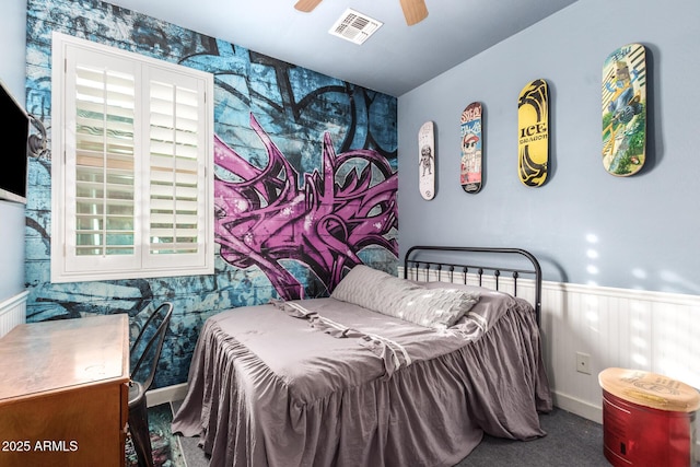 carpeted bedroom with a wainscoted wall, multiple windows, visible vents, and a ceiling fan