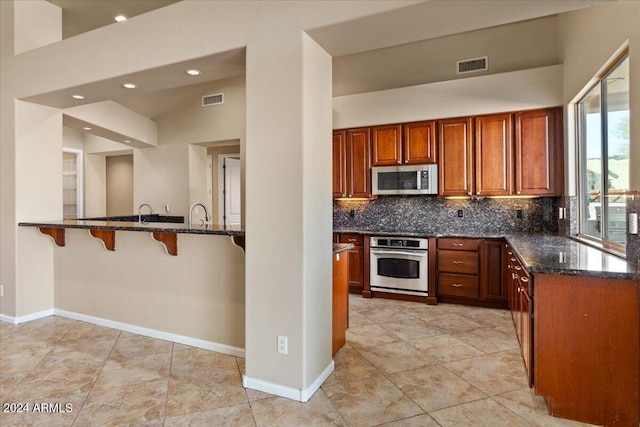 kitchen featuring stainless steel appliances, backsplash, kitchen peninsula, dark stone counters, and a breakfast bar