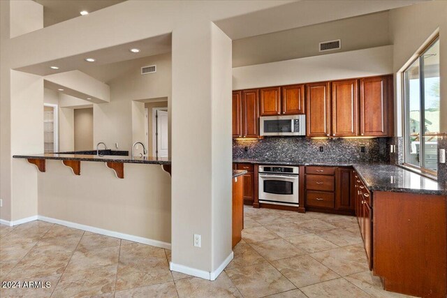 36908 N 19th St, Phoenix, AZ 85086 - kitchen with stainless steel appliances, dark stone counters, brown cabinetry, backsplash, and a breakfast bar