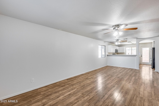 unfurnished living room with a ceiling fan, wood finished floors, baseboards, arched walkways, and a textured ceiling