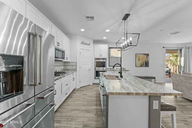 kitchen with a kitchen island with sink, white cabinets, sink, light stone countertops, and stainless steel appliances