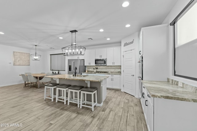 kitchen with white cabinets, light stone countertops, an island with sink, appliances with stainless steel finishes, and decorative light fixtures