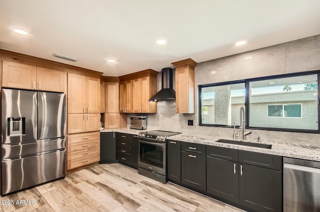 kitchen with appliances with stainless steel finishes, light stone counters, wall chimney exhaust hood, sink, and light hardwood / wood-style flooring