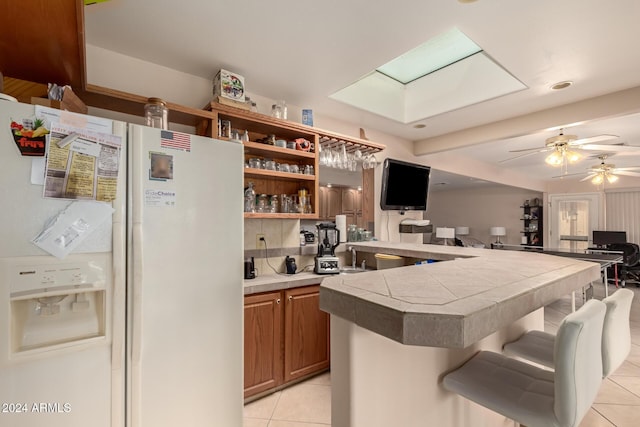 kitchen with a breakfast bar, ceiling fan, light tile patterned floors, tile countertops, and white fridge with ice dispenser