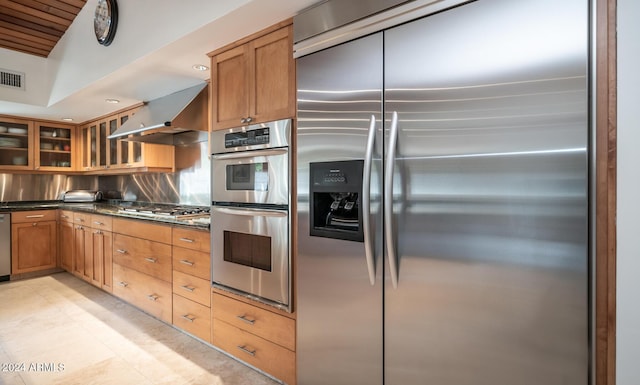 kitchen featuring backsplash, dark stone counters, wall chimney range hood, light tile patterned floors, and appliances with stainless steel finishes