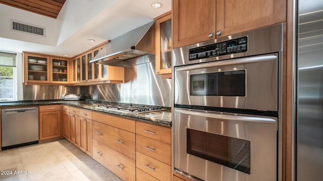 kitchen featuring tasteful backsplash, dark stone counters, stainless steel appliances, wall chimney range hood, and light tile patterned flooring