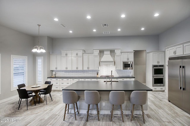 kitchen with a center island with sink, dark countertops, decorative light fixtures, built in appliances, and custom exhaust hood