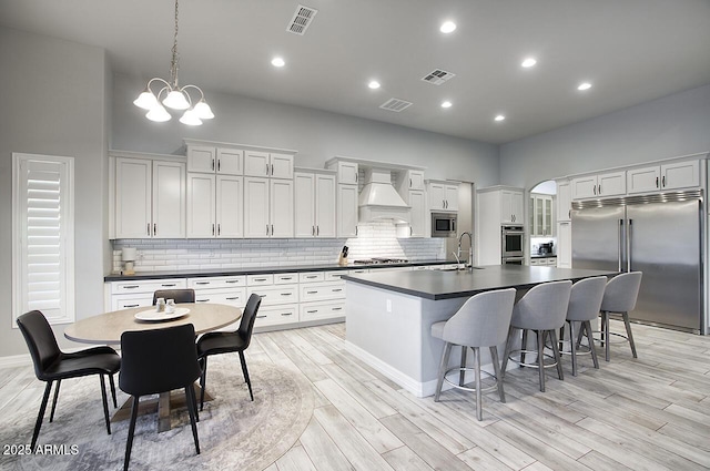 kitchen with dark countertops, custom range hood, white cabinets, an island with sink, and built in appliances