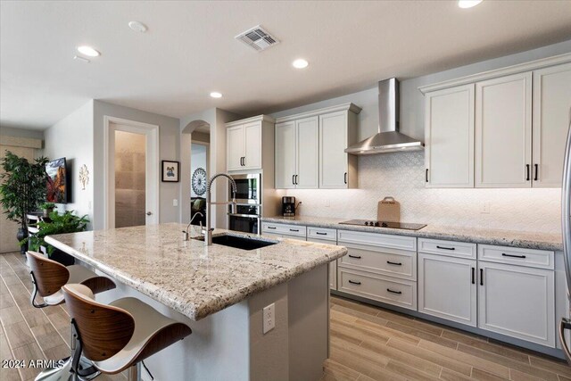 kitchen with arched walkways, an island with sink, wall chimney range hood, white cabinetry, and a sink