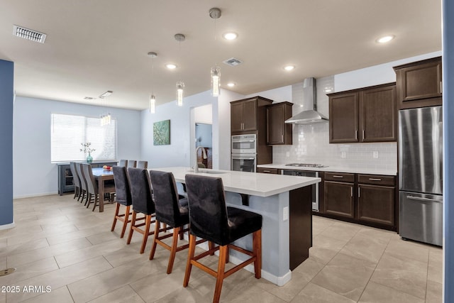 kitchen with a kitchen bar, stainless steel appliances, a kitchen island with sink, wall chimney range hood, and hanging light fixtures