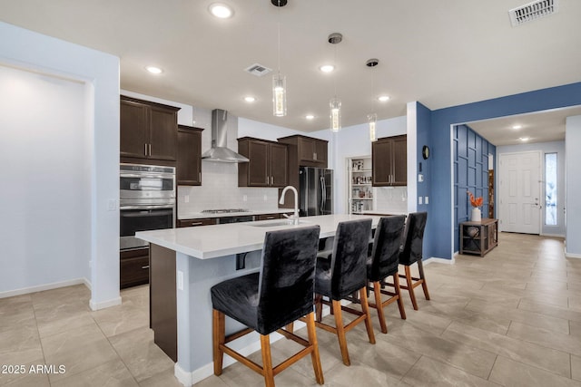 kitchen with sink, wall chimney exhaust hood, hanging light fixtures, an island with sink, and appliances with stainless steel finishes