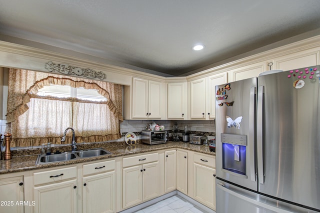 kitchen with stainless steel refrigerator with ice dispenser, cream cabinetry, sink, and dark stone countertops