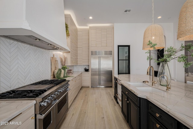 kitchen with sink, high end appliances, custom range hood, hanging light fixtures, and light stone countertops