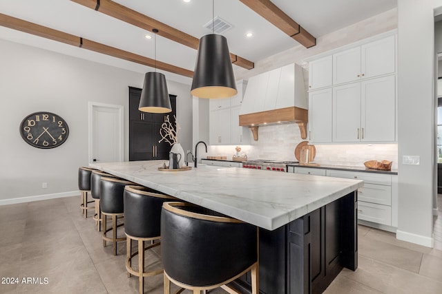 kitchen featuring beamed ceiling, premium range hood, white cabinets, and an island with sink