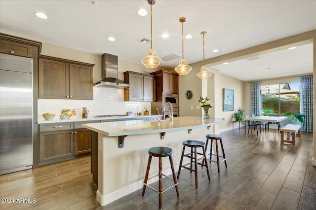 kitchen with pendant lighting, wall chimney exhaust hood, dark hardwood / wood-style floors, an island with sink, and appliances with stainless steel finishes