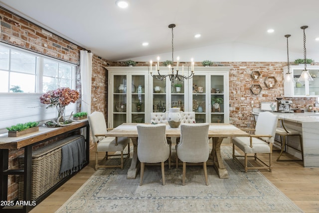 dining space with brick wall, light hardwood / wood-style floors, vaulted ceiling, and a notable chandelier