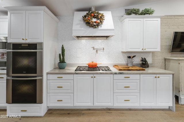 kitchen with appliances with stainless steel finishes, white cabinetry, backsplash, light hardwood / wood-style floors, and custom range hood