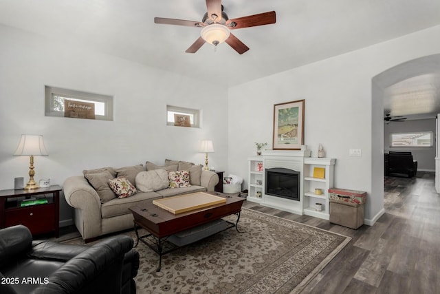living area with arched walkways, ceiling fan, wood finished floors, baseboards, and a glass covered fireplace