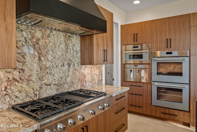 kitchen with light stone countertops, decorative backsplash, appliances with stainless steel finishes, wall chimney exhaust hood, and modern cabinets