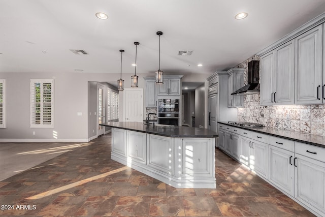 kitchen with a center island with sink, stainless steel gas stovetop, wall chimney exhaust hood, hanging light fixtures, and double oven