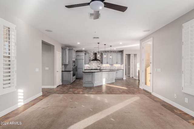 kitchen with wall chimney exhaust hood, a kitchen island with sink, gray cabinets, backsplash, and decorative light fixtures