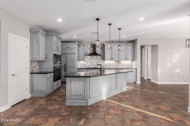 kitchen featuring tasteful backsplash, decorative light fixtures, an island with sink, gray cabinets, and wall chimney range hood