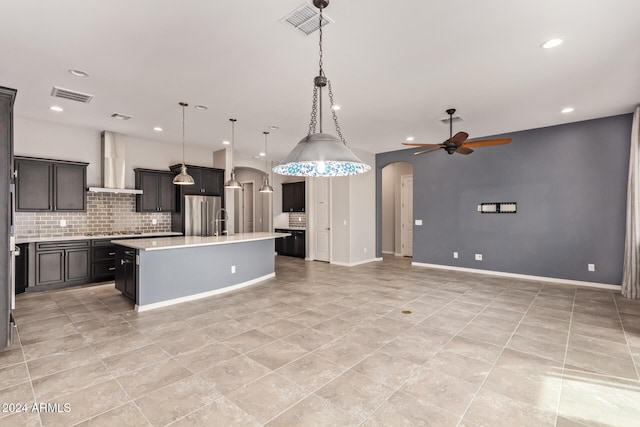 kitchen with appliances with stainless steel finishes, wall chimney exhaust hood, ceiling fan, a kitchen island with sink, and pendant lighting