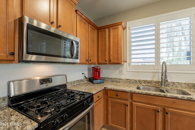 kitchen with appliances with stainless steel finishes, a sink, backsplash, brown cabinetry, and light stone counters
