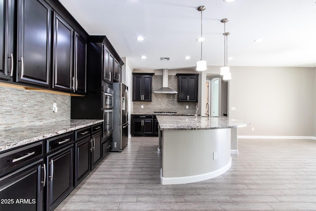 kitchen featuring tasteful backsplash, an island with sink, hanging light fixtures, light stone countertops, and wall chimney range hood