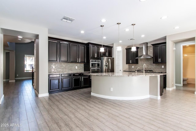 kitchen with decorative backsplash, hanging light fixtures, stainless steel appliances, a center island with sink, and wall chimney range hood