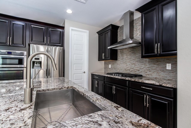 kitchen with sink, tasteful backsplash, stainless steel appliances, light stone countertops, and wall chimney range hood