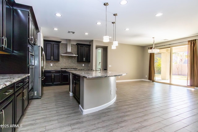 kitchen with wall chimney exhaust hood, tasteful backsplash, gas cooktop, pendant lighting, and a kitchen island with sink