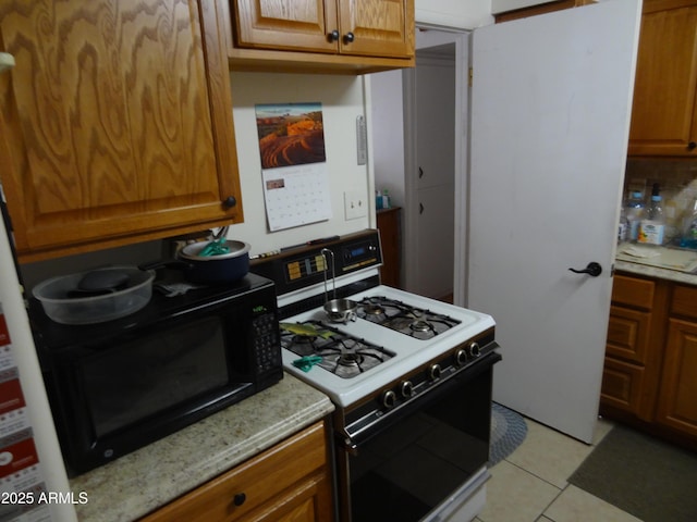 kitchen with black microwave, light tile patterned floors, light stone counters, brown cabinetry, and gas range oven