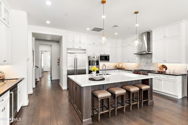 kitchen with a center island with sink, dark countertops, built in appliances, decorative light fixtures, and wall chimney range hood