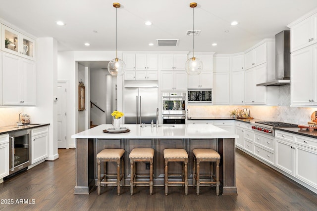 kitchen with dark countertops, wine cooler, appliances with stainless steel finishes, hanging light fixtures, and wall chimney range hood