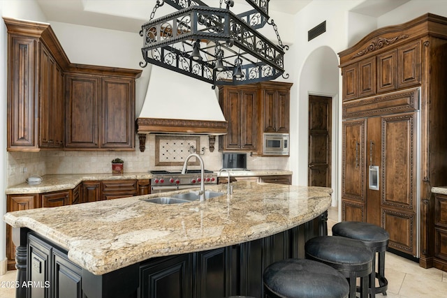 kitchen with custom exhaust hood, a center island with sink, backsplash, built in appliances, and sink