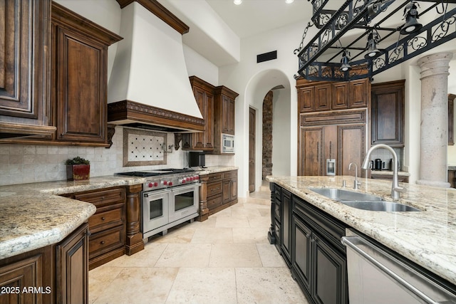 kitchen with premium appliances, custom range hood, light stone countertops, dark brown cabinetry, and sink