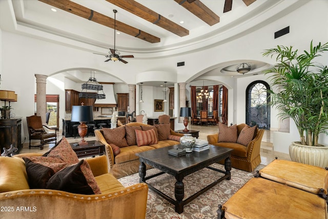 living room featuring ceiling fan with notable chandelier, beam ceiling, ornate columns, and a towering ceiling